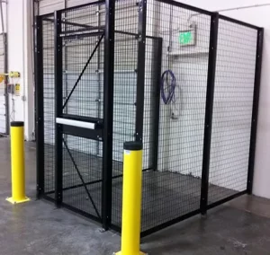 A secure wire mesh cage with a locked door, designed for Warehouse Solutions, is located indoors near a wall with an exit sign and two yellow protective bollards in front.