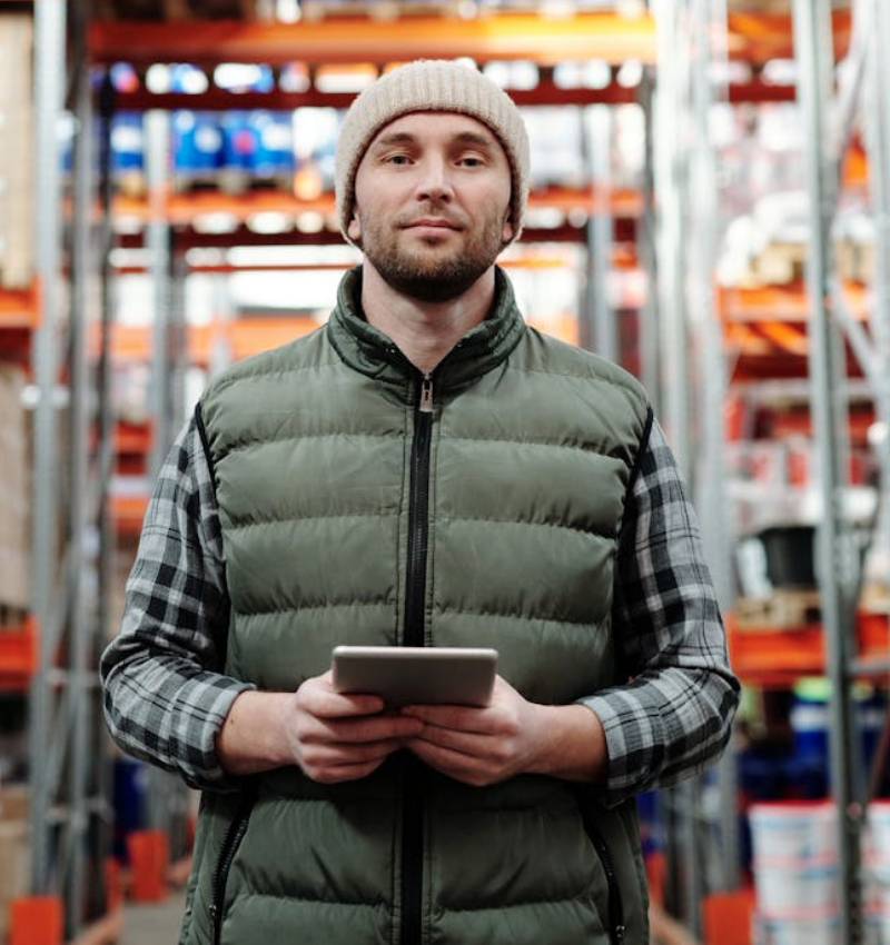 A man wearing a green vest and beanie holds a tablet while standing in a warehouse aisle, showcasing efficient Warehouse Solutions with shelves in the background.