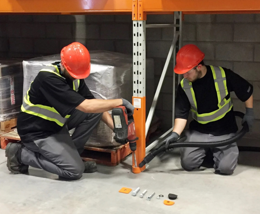 Two workers in safety gear drill into a warehouse floor beside pallet racks, with tools and fasteners laid out in front of them—demonstrating efficient Warehouse Solutions in action.