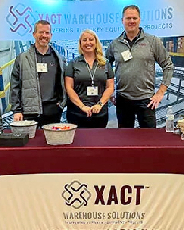 Three people stand behind a table with XACT Warehouse Solutions branding at a trade show booth, smiling for the camera and ready to share more about Xact Warehouse Solutions.