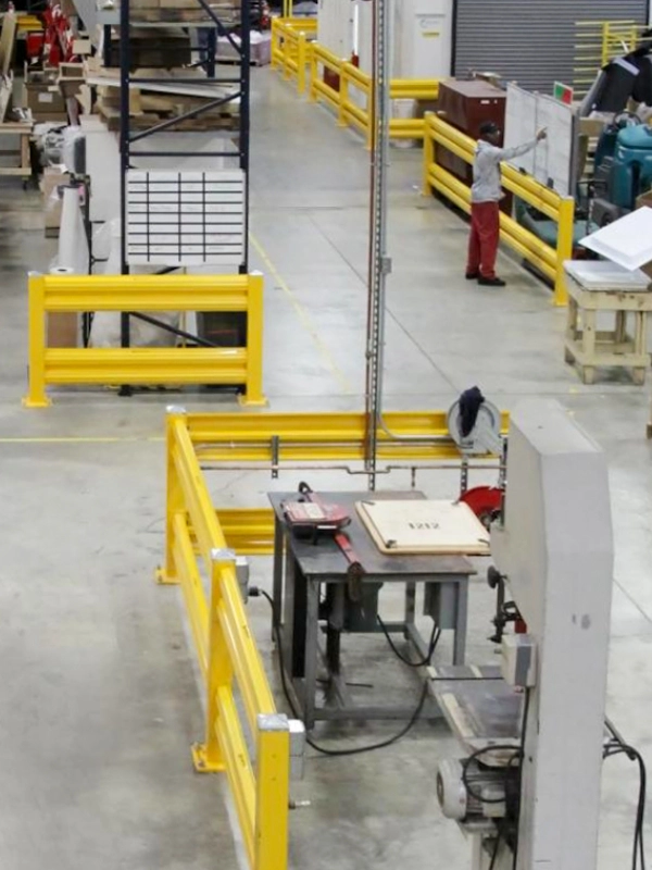 Industrial workshop featuring yellow safety barriers and industrial warehouse safety guarding, with machinery, work tables, and one person standing near equipment in the background.
