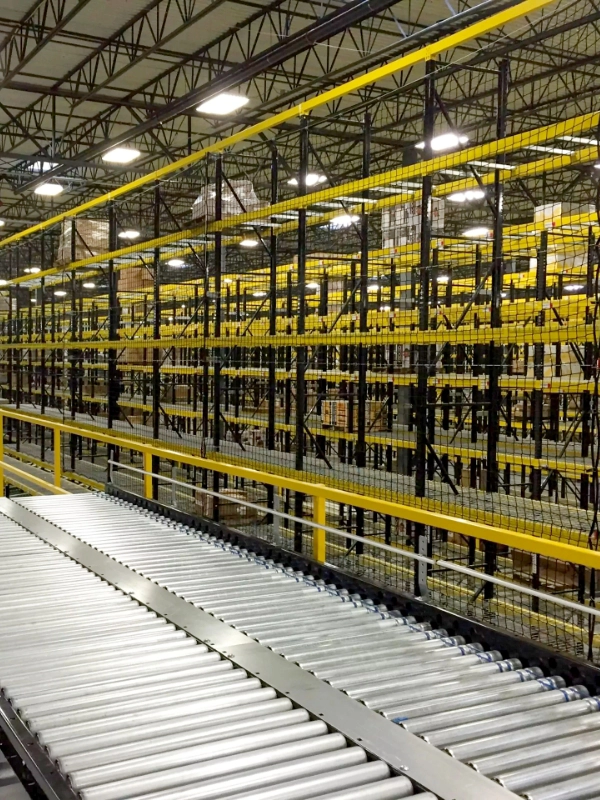 A warehouse interior with empty metal storage racks, yellow railings, Warehouse Safety Netting, and a metal roller conveyor in the foreground.