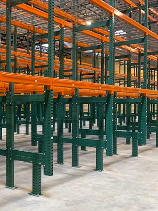 Rows of green and orange Selective Rack steel warehouse storage racks stand empty on a concrete floor inside a large industrial building.