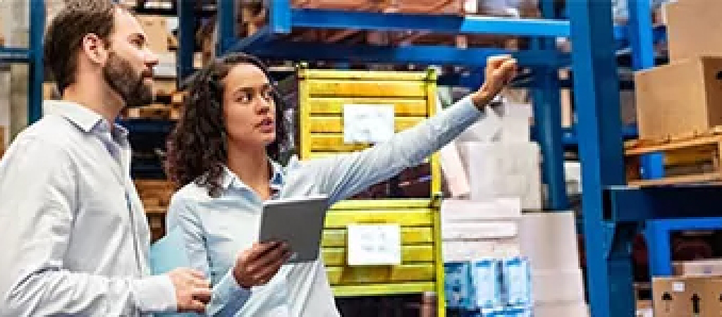 Two people in business attire stand in a warehouse; one holds a tablet and gestures toward shelves stacked with boxes while the other listens, discussing the details of Turnkey Warehouse Projects.