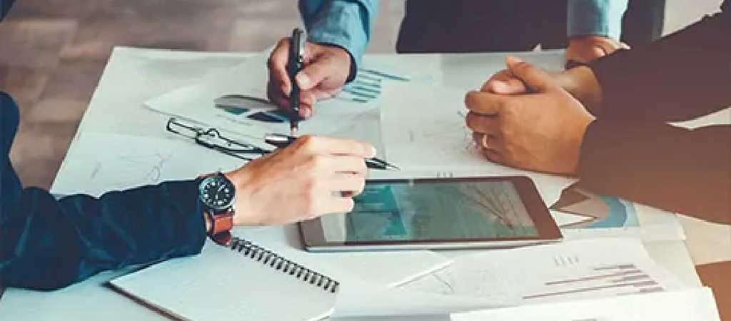 Three people are gathered around a table with documents, a notepad, and a tablet, reviewing papers and discussing plans for Turnkey Warehouse Projects during their meeting.