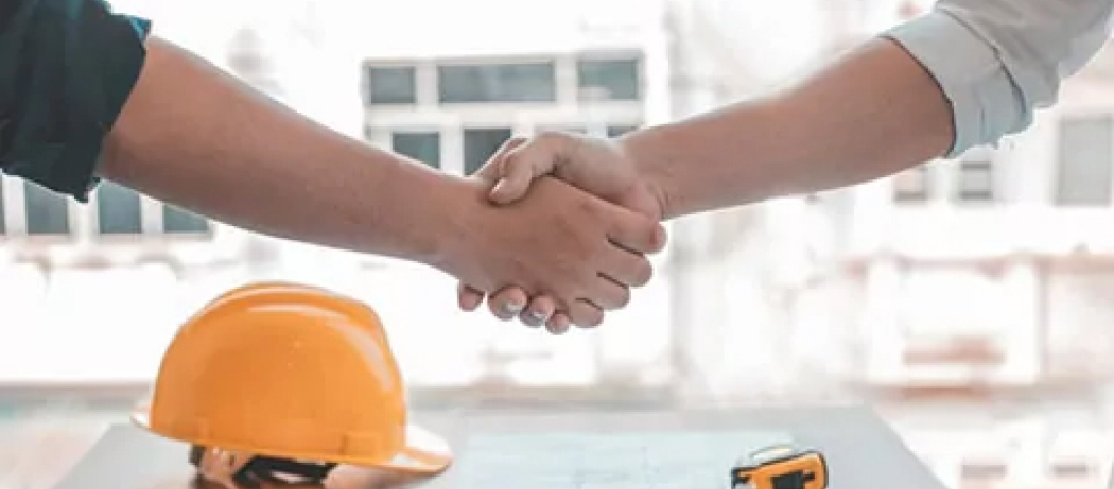 Two people shaking hands over a table with a yellow construction helmet, a blueprint, and a tape measure—sealing the deal on Turnkey Warehouse Projects.