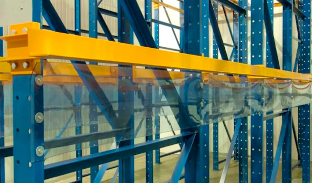 Blue and yellow industrial Drive-In Rack storage racks are bolted together in a warehouse, with transparent plastic safety barriers attached to the lower section.