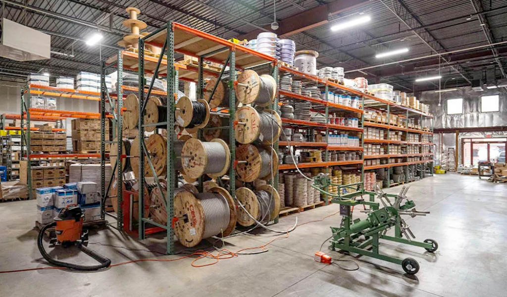 Industrial warehouse interior illustrating the diverse Industries We Serve, with shelves holding large spools of cable, boxes, and supplies; machinery and equipment are visible in the foreground.
