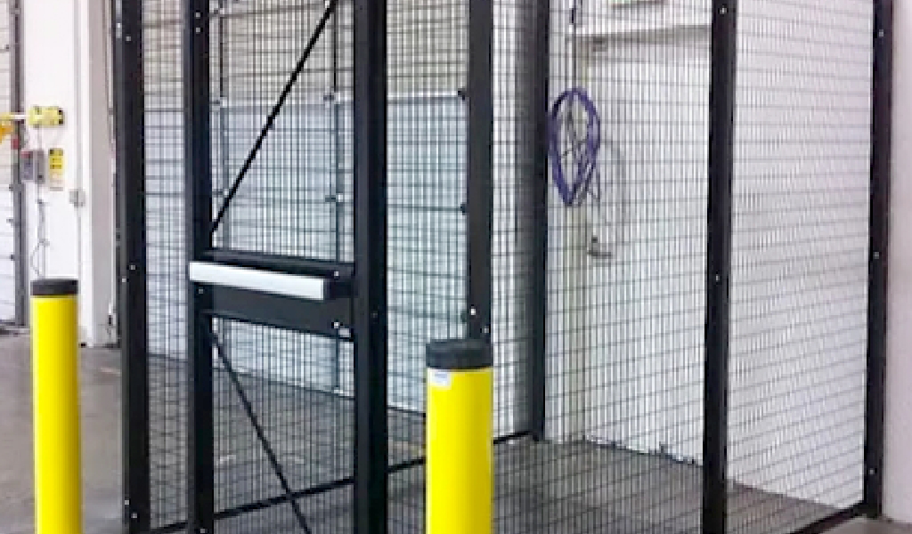 A metal security cage with mesh panels, a door with a push bar, and yellow safety bollards serves as reliable industrial safety guard fencing in an indoor setting.
