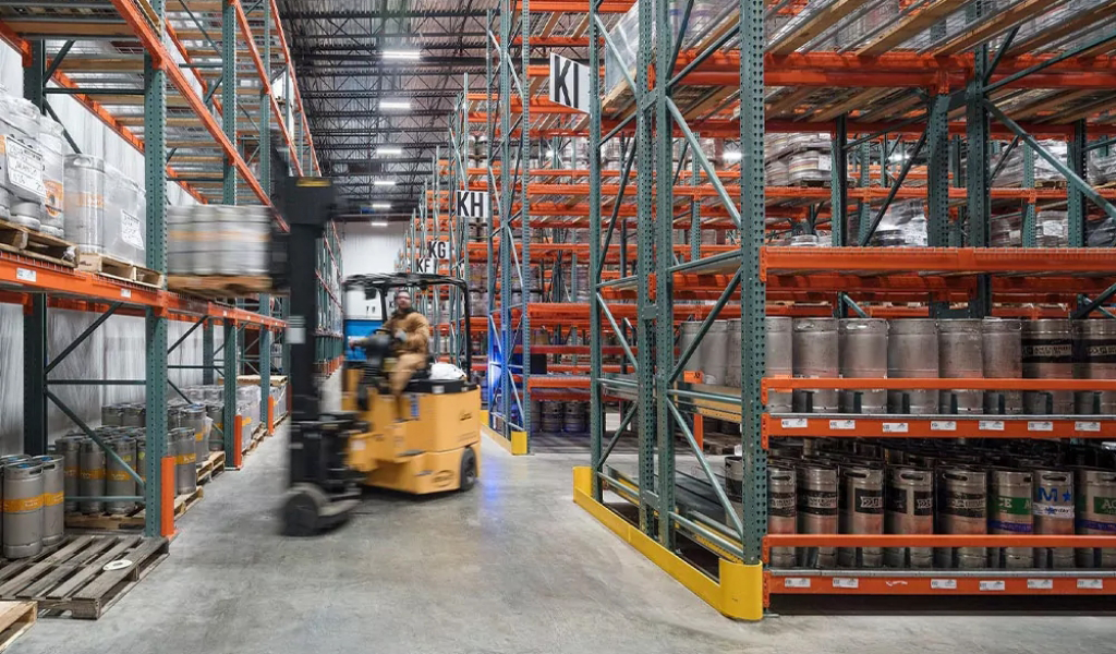 A warehouse interior with metal shelves holding industrial barrels and kegs; a person drives a forklift down the aisle, showcasing the dynamic environments within the Industries We Serve.