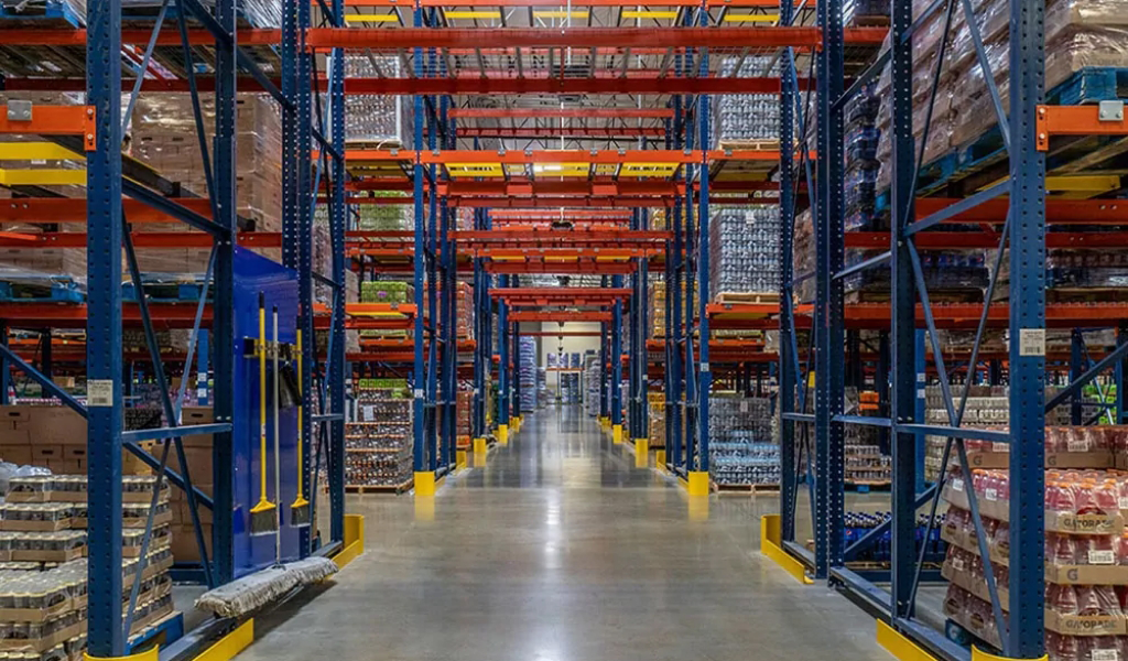 Tall shelves stocked with various goods line both sides of a wide, empty aisle inside a brightly lit industrial warehouse, reflecting the diverse needs of the industries we serve.