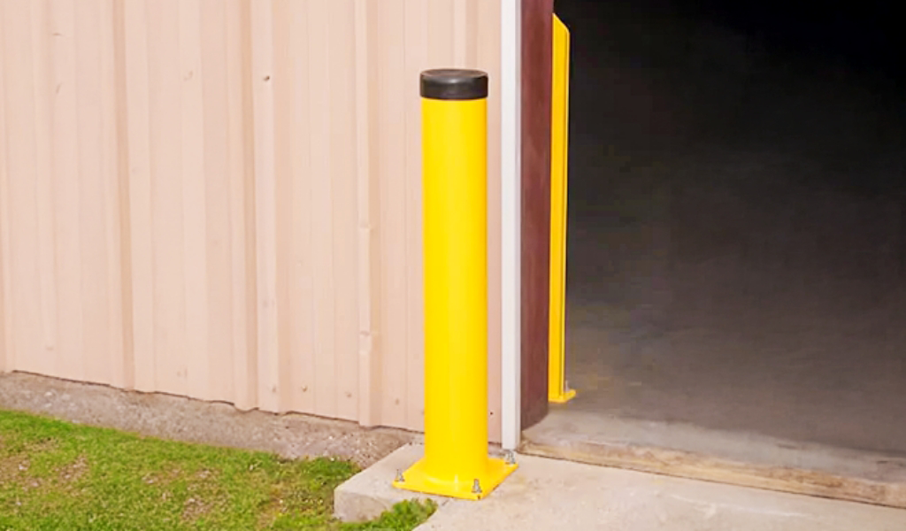 A yellow safety bollard is installed near the corner of a beige metal building next to an open doorway, providing essential industrial warehouse safety guarding.
