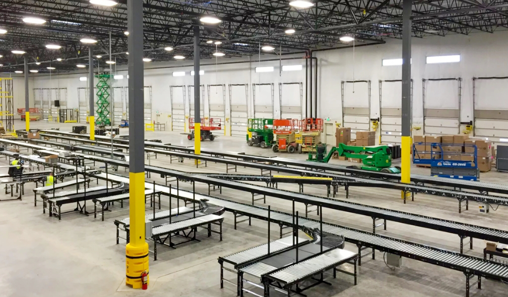 Large warehouse interior with empty conveyor belts, high ceilings, industrial lighting, and various equipment and boxes arranged near loading docks featuring Industrial Warehouse Safety Guarding.