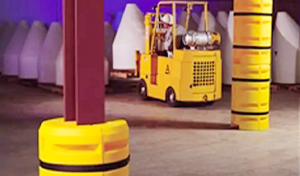 A person operates a yellow forklift in an industrial warehouse, maneuvering between padded columns and white objects covered with tarps, demonstrating effective warehouse safety guarding.