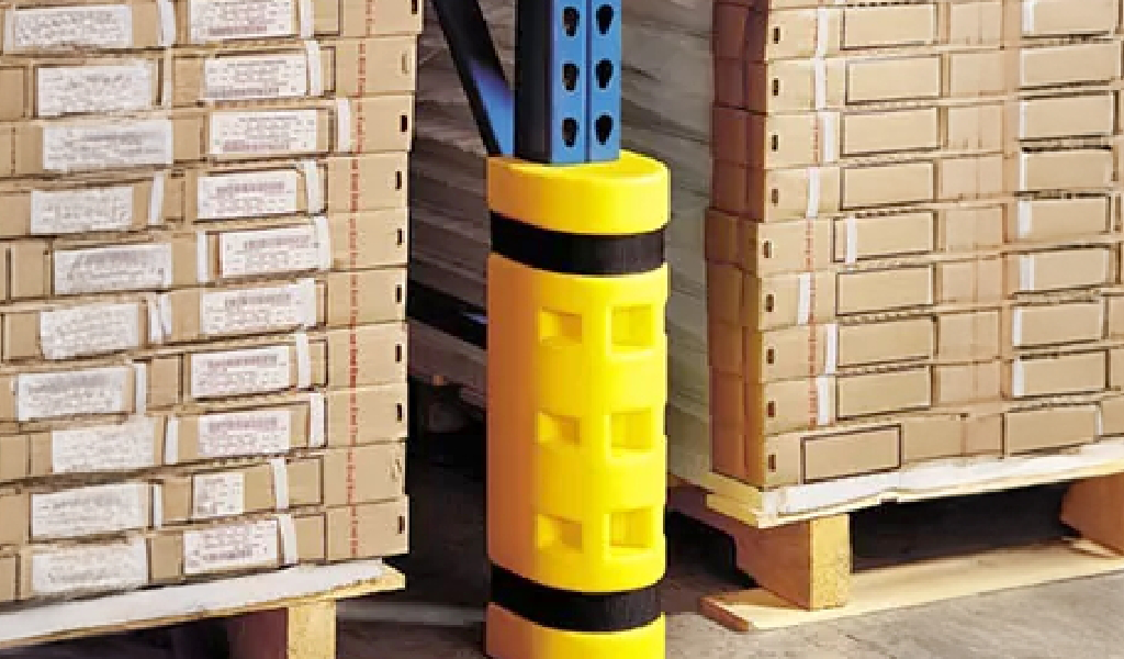 A yellow industrial warehouse safety guarding bumper is wrapped around a blue warehouse rack post between stacked pallets of cardboard boxes on wooden pallets.