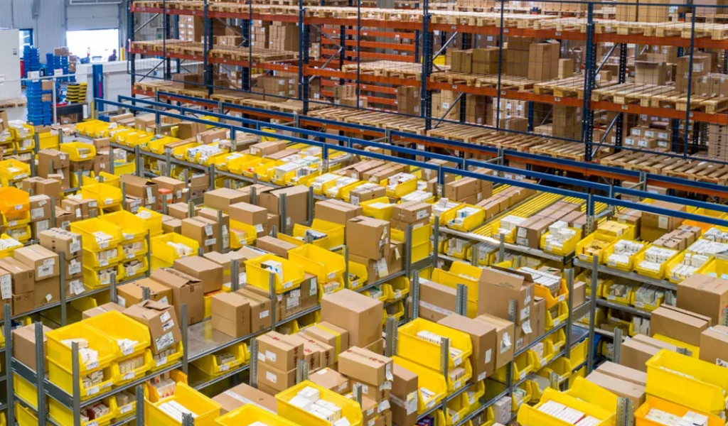 Rows of metal shelves filled with yellow bins and cardboard boxes are organized in a warehouse storage area, showcasing the efficient storage solutions provided for the diverse Industries We Serve.