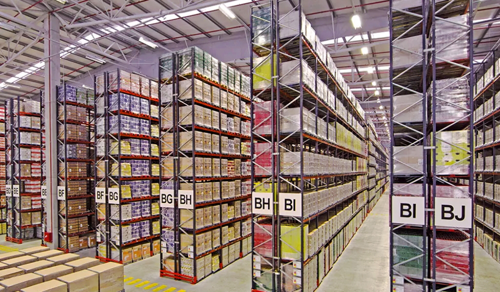 Large warehouse interior with tall selective rack shelves filled with stacked boxes and labeled sections, including signs for BG, BH, and BI. Bright lighting and an organized storage layout complete the efficient space.
