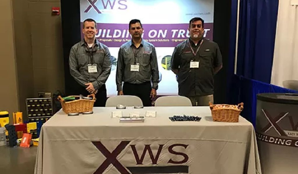 Three men stand behind a table with promotional materials at an XWS trade show booth. A banner reading “BUILDING ON TRUST” hangs in the background, highlighting what’s special about Xact Warehouse Solutions.