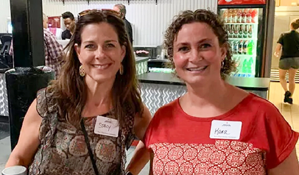 Two women wearing name tags stand side by side and smile in front of a drink cooler in a casual indoor setting, representing About Xact Warehouse Solutions with friendly professionalism.