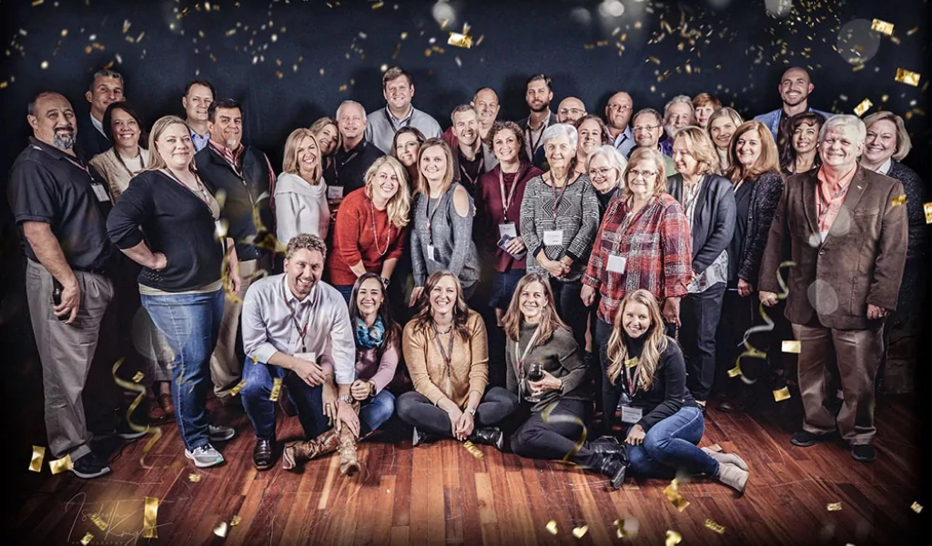 A large group of adults, standing and sitting on a wooden floor, pose for a group photo indoors with gold confetti falling around them—celebrating another milestone at About Xact Warehouse Solutions.
