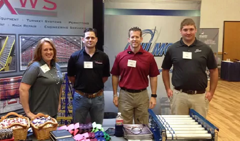 Four people stand behind a display booth at a trade show for Xact Warehouse Solutions, featuring equipment brochures, baskets with items, and a conveyor roller demo on the table.