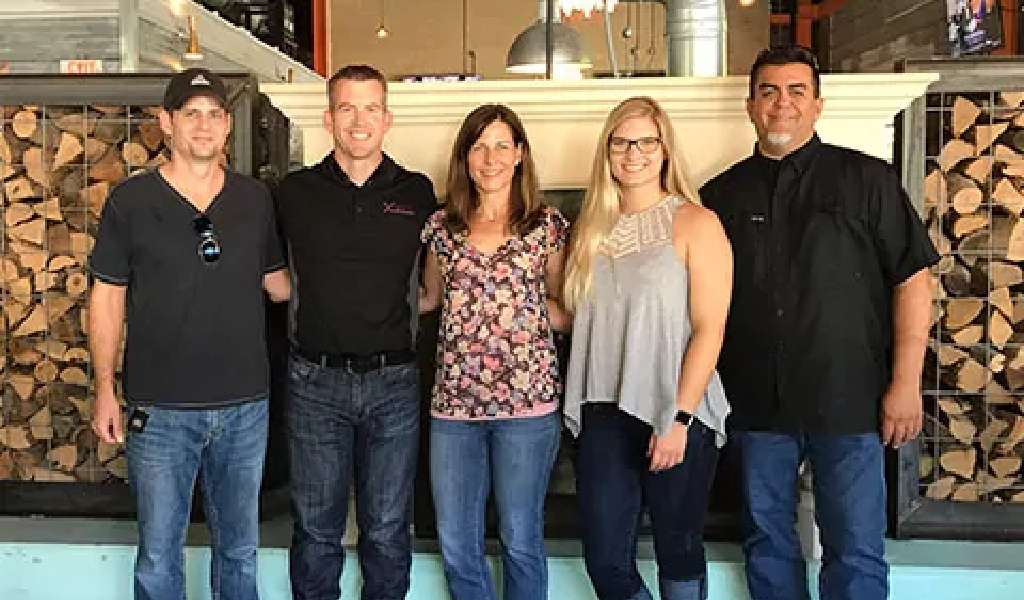 Five people stand side by side indoors in front of a stacked wood display, posing and smiling for the photo—capturing a team moment at About Xact Warehouse Solutions.