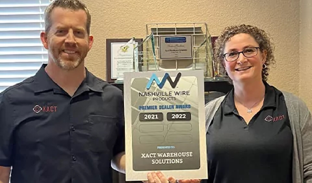 Two people in matching XACT shirts hold a framed Nashville Wire Products Premier Dealer Award for XACT Warehouse Solutions, with years 2021 and 2022 visible—a proud moment that says a lot about Xact Warehouse Solutions.