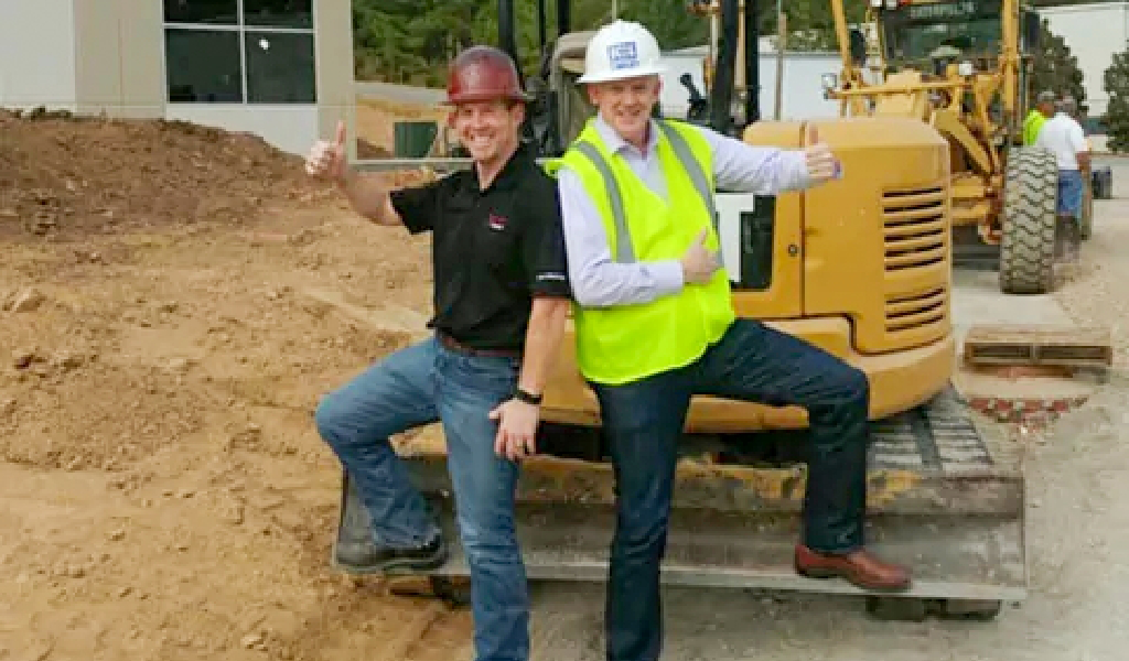 Two men wearing hard hats and business attire pose with a thumbs-up gesture in front of construction equipment at a work site, showcasing the professionalism found at About Xact Warehouse Solutions.