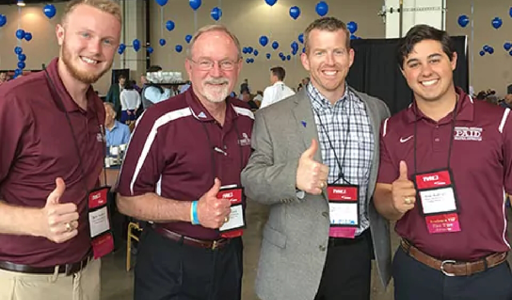 Four men stand indoors at an event for Xact Warehouse Solutions, three in maroon shirts and one in a checked shirt. All are smiling and giving thumbs up, with blue balloons visible in the background.