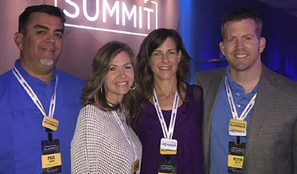 Four people wearing name badges stand together and smile at a summit event, with a sign reading "SUMMIT" in the background, representing About Xact Warehouse Solutions.