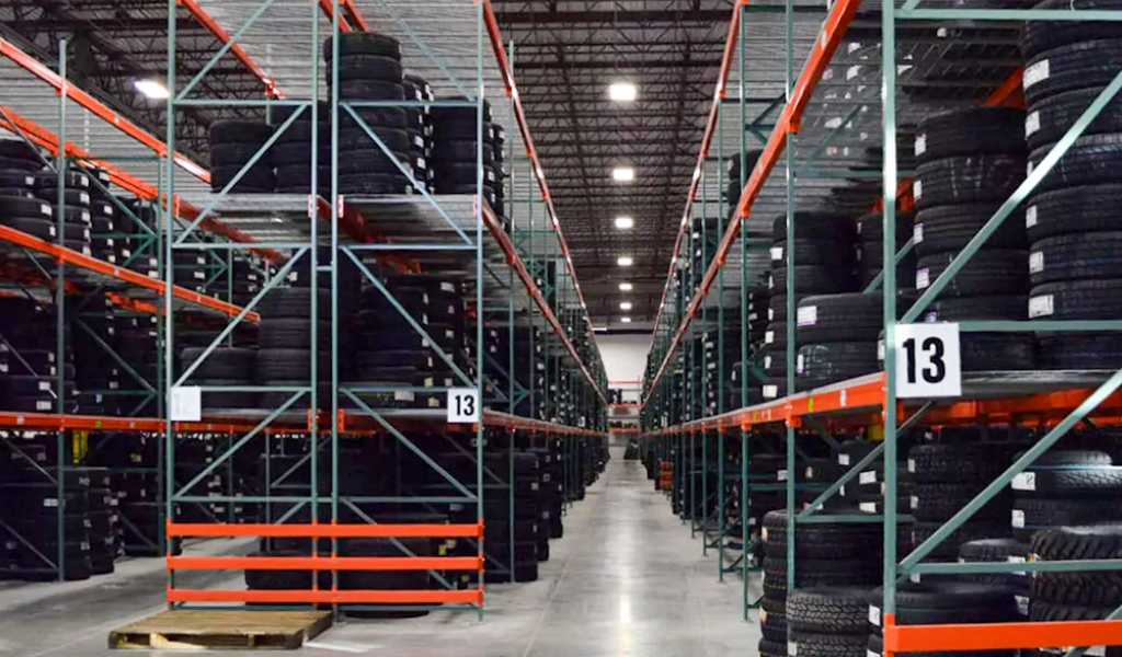 Warehouse interior with tall metal racks holding stacked tires, organized in rows with numbered aisle signs, under a high ceiling with industrial lighting—ideal for the diverse needs of the Industries We Serve.
