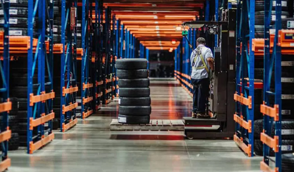 A warehouse worker operates a forklift beside stacked tires on racks, framed by rows of orange and blue shelving—just one example from the many Industries We Serve.
