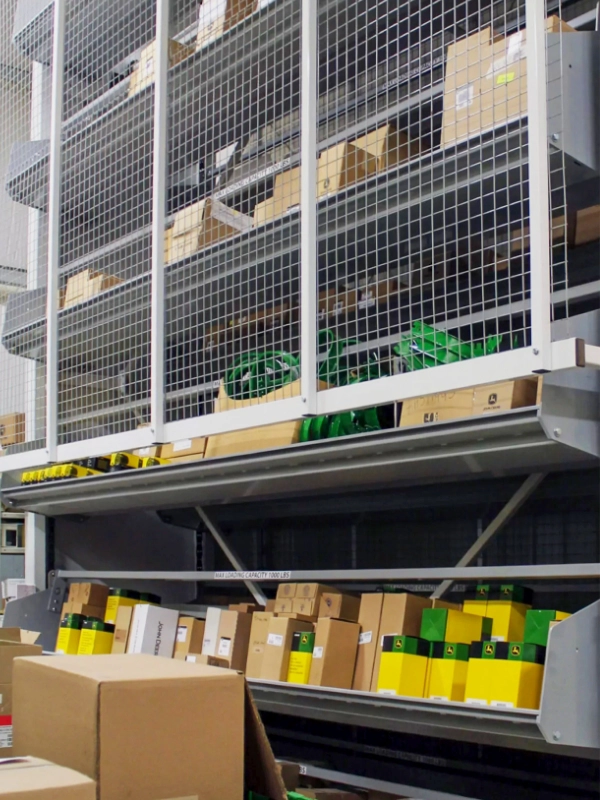 Metal shelving units with wire mesh barriers hold numerous cardboard boxes and packages, some labeled in green and yellow, alongside vertical carousels for efficient organization in the warehouse storage area.