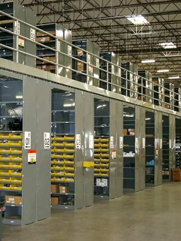 Rows of industrial shelving units in a warehouse, with labeled bins and shelves filled with various items, and a mezzanine level above.