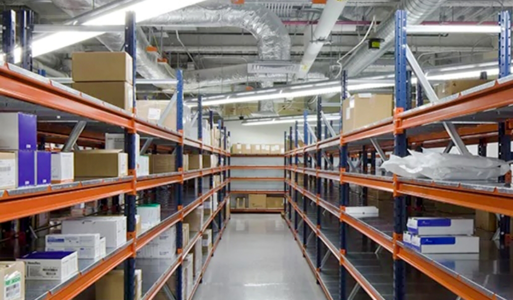 Warehouse shelves stacked with various boxes and packages under fluorescent lighting, featuring industrial shelving and visible ceiling pipes and ductwork above.