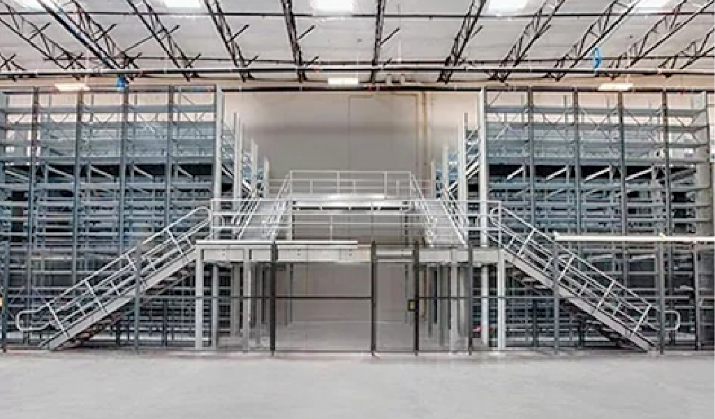 A large industrial warehouse interior featuring industrial shelving units and two wide staircases leading to an upper platform.