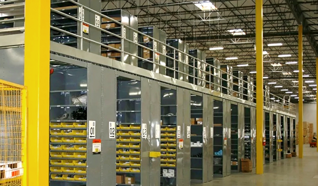 Warehouse interior with industrial shelving units, labeled bins, and a mezzanine level under high ceilings, all illuminated by overhead lights.