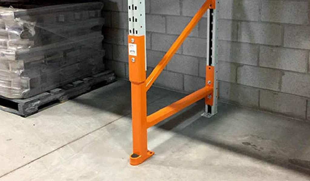 Orange steel rack guard installed at the base of a warehouse shelving unit near a brick wall and a pallet wrapped in plastic, offering added protection as part of comprehensive rack repair kits.